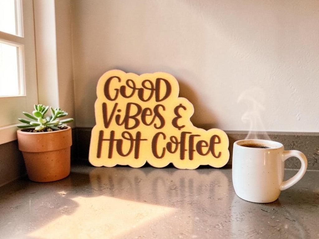 permanent vinyl sticker with 'Good Vibes & Hot Coffee' text on a countertop next to a steaming mug and plant