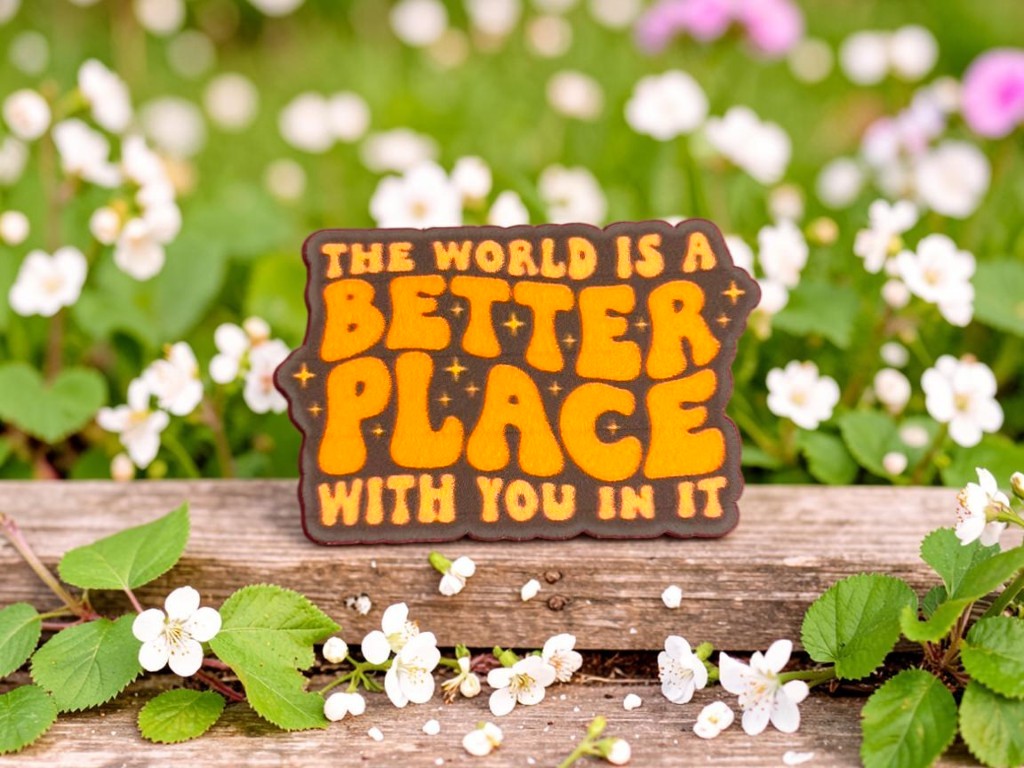 Permanent Sticker with 'The world is a better place with you in it' text on a wooden log with flowers in the background